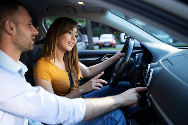 A driving instructor guides a student through a pre-drive safety check, pointing to the car's tires