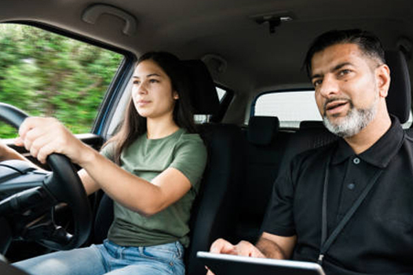 A driving instructor with dual controls looks attentively at the road ahead while a student practices navigating a busy intersection.