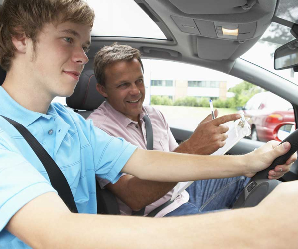 From the driver's point of view, the hands of a learner grip the steering wheel with a driving instructor visible beside them.