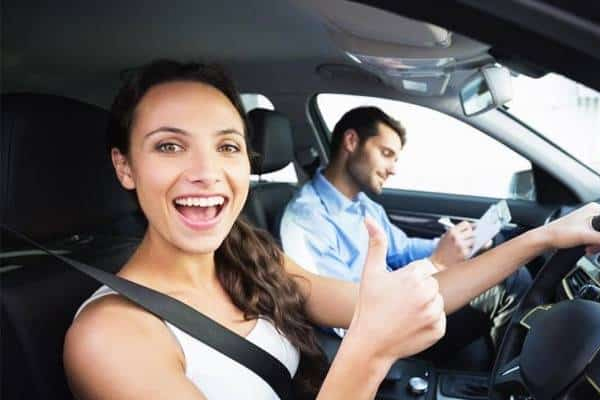 A driving instructor points to a road sign while providing instructions to a young learner driver from the passenger seat.