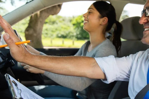 A driving instructor points to a road sign while providing instructions to a young learner driver from the passenger seat.