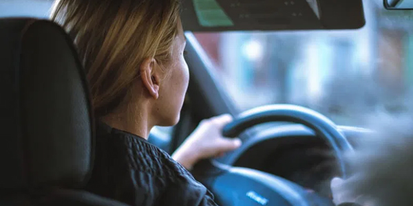 A student driver carefully practices parallel parking a white driving school car between orange cones in a large, empty lot.