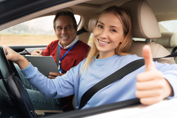 A driving instructor calmly gestures while providing guidance to a focused student driver from the passenger seat.